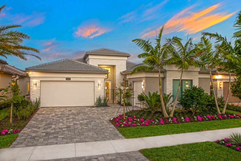Modern single-story house with a paved driveway, two-car garage, manicured lawn, vibrant flowerbeds, palm trees, and exterior lighting—an Itzhak-inspired design set against a colorful evening sky with pink and blue hues. | FintechZoom