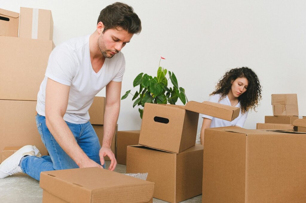 A man and woman wearing casual clothes pack cardboard boxes in a bright room, with a green potted plant and several moving boxes around them, suggesting they are preparing for a long-distance move or just arriving. | FintechZoom