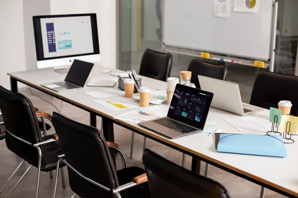 A conference room table with open laptops, paper documents, coffee cups, and stationery hints at an ongoing IT procurement meeting. Empty chairs surround the table, while a large monitor displays graphs and charts. A whiteboard is visible in the background. | FintechZoom