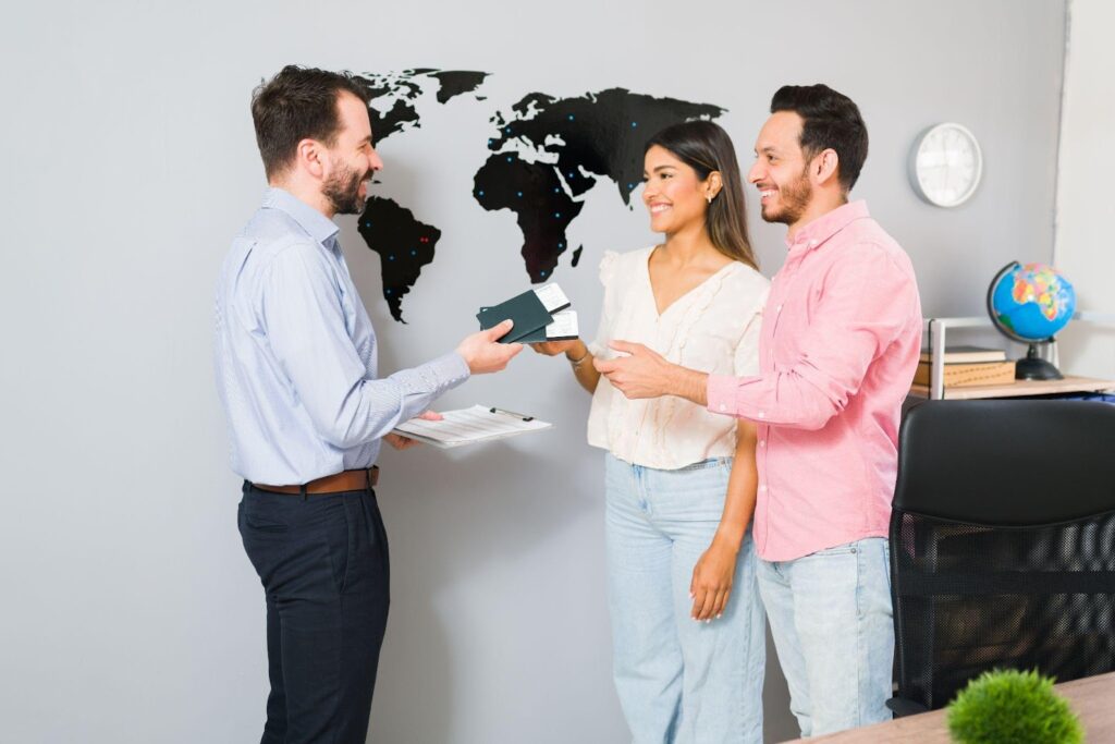 A man hands passports, flight tickets, and travel insurance documents to a smiling couple in an office with a world map on the wall, suggesting a travel agency or trip planning setting. | FintechZoom