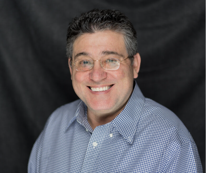 A smiling man named Itzhak with short, curly dark hair and glasses, wearing a blue and white checkered shirt, poses in front of a dark, plain background. | FintechZoom