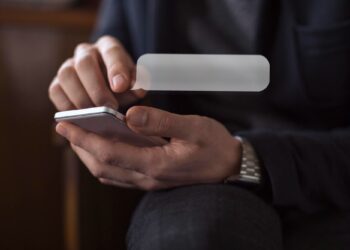 A person wearing a dark jacket is using a smartphone, with one hand holding the phone and the other hand touching the screen. A transparent chat bubble is digitally superimposed above the phone. | FintechZoom