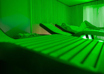 A person in a towel reclines on a lounge chair in a dimly lit room with green lighting and vertical striped walls, creating a relaxing spa-like atmosphere. | FintechZoom