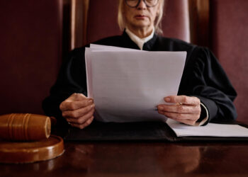 A judge in a black robe sits at a desk, holding and reading documents. A wooden gavel and a folder rest on the desk in front of her. The judge’s face is partially visible. | FintechZoom