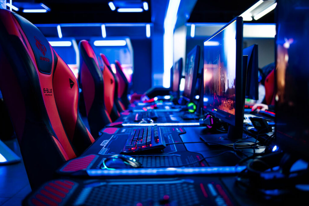 A row of gaming PCs with illuminated keyboards and red-and-black gaming chairs lines a dark room, blue LED lights casting a futuristic gaming atmosphere. | FintechZoom