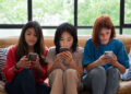 Three young women sit on a couch, each focused on their smartphone. They appear relaxed, with greenery visible through large windows behind them. | FintechZoom