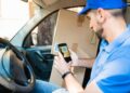 A delivery driver in a blue shirt and cap sits in a vehicle, using his phone to scan a QR code on a large cardboard box placed beside him. | FintechZoom