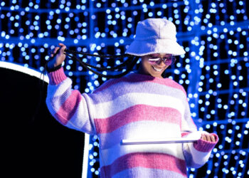 A person wearing a bucket hat, sunglasses, and a pink-and-white striped sweater smiles while holding a tablet. Blue and white string lights create a bright, festive background. | FintechZoom