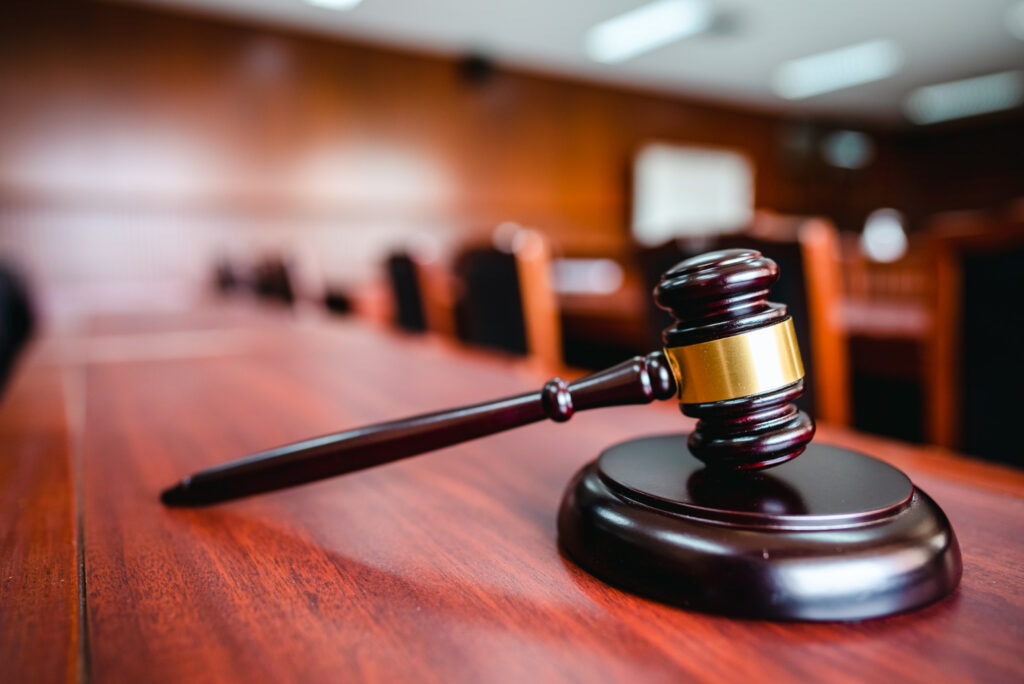 A wooden judge’s gavel rests on a sound block on a polished courtroom table, with rows of empty chairs and blurred wood-paneled walls in the background. | FintechZoom