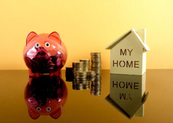 A pink piggy bank, stacks of coins, and a small wooden house with "MY HOME" written on it are arranged against a yellow background, all reflected on a glossy surface. | FintechZoom