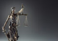 A bronze statue of Lady Justice holding balanced scales in one hand and a sword in the other, wearing a blindfold, set against a dark gradient background. | FintechZoom