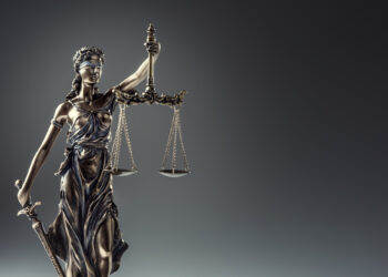 A bronze statue of Lady Justice holding balanced scales in one hand and a sword in the other, wearing a blindfold, set against a dark gradient background. | FintechZoom