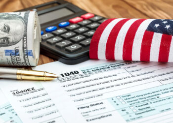 A rolled-up $100 bill, a calculator, an American flag, and a pen sit on top of U.S. tax forms, including a 1040 form, representing tax preparation and finance. | FintechZoom