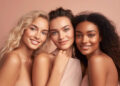 Three young women with different skin tones and hair types smile closely together against a soft peach background, showcasing natural beauty and glowing, healthy skin. | FintechZoom