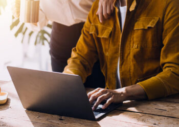 A person in a mustard jacket types on a laptop at a wooden table while another person stands beside them, holding a mug and resting a hand on their shoulder in a bright room. | FintechZoom