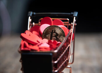 A small shopping cart filled with red heart-shaped tokens and a single gold Bitcoin coin, placed on a wooden surface against a dark background. | FintechZoom