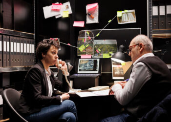 Two people sit at a desk in a dimly lit office, discussing a case. Behind them are crime scene photos, notes, and evidence connected with string on a wall, suggesting an investigation or detective work. | FintechZoom
