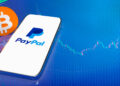 A smartphone displays the PayPal logo on its screen, next to a large orange Bitcoin symbol, set against a blue digital background with financial graph lines. | FintechZoom