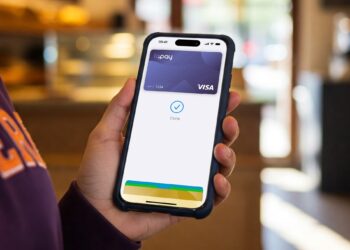 A person holds a smartphone showing a digital wallet app with a VISA card, displaying a check mark and “Done” on the screen, indicating a successful payment in a cafe or restaurant setting. | FintechZoom