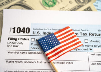 A small American flag on a toothpick lies on top of a U.S. 1040 income tax return form, with part of a dollar bill visible in the background. | FintechZoom