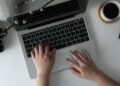 A person types on a laptop at a white desk with Vidnoz AI open, surrounded by a camera, tablet, books, coffee, and colored markers—a setting that suggests a creative or work-from-home environment. | FintechZoom