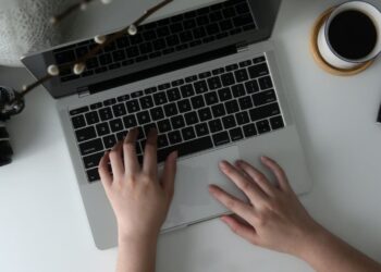 A person types on a laptop at a white desk with Vidnoz AI open, surrounded by a camera, tablet, books, coffee, and colored markers—a setting that suggests a creative or work-from-home environment. | FintechZoom