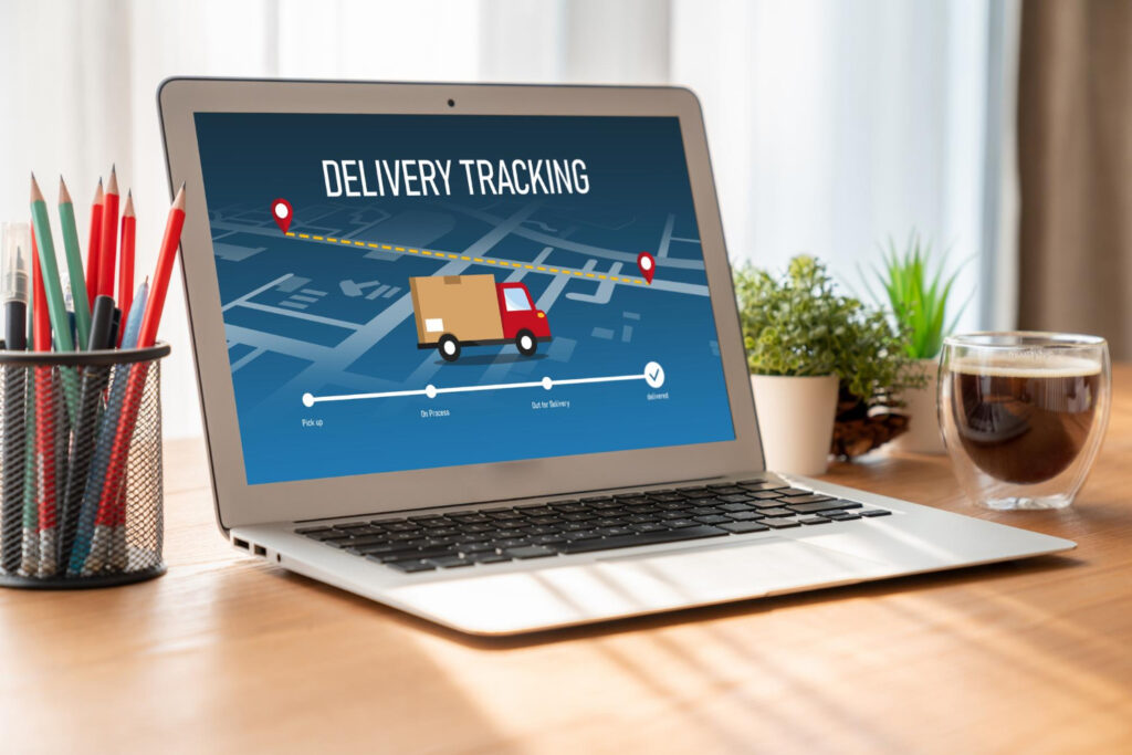 A laptop on a wooden desk displays a delivery tracking screen with a map, route, and a moving truck. Nearby are colored pencils in a holder, a small plant, and a glass cup of coffee. | FintechZoom