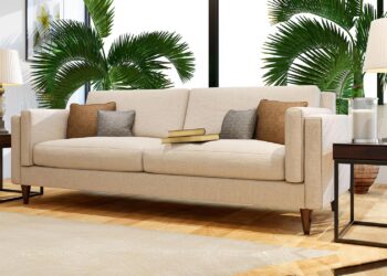 A modern living room with a beige sofa, four throw pillows, two books on the seat, dark wood side tables with lamps, large green plants, and sunlight streaming through tall windows. | FintechZoom