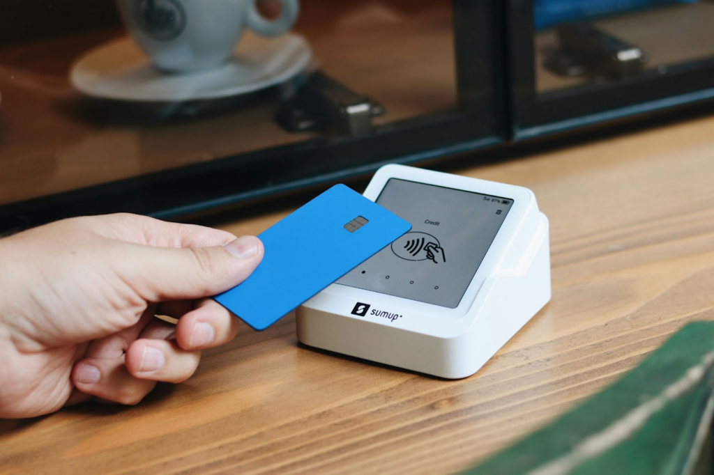 A person holds a blue contactless card near a white SumUp card reader on a wooden table, preparing to make a payment. A coffee cup and glass divider are visible in the background. | FintechZoom