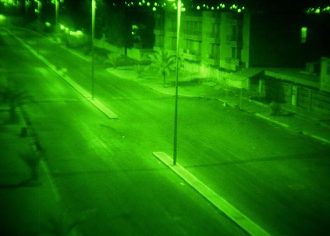A deserted urban street at night viewed through a green night vision filter, with streetlights, empty sidewalks, and buildings lining the road. | FintechZoom