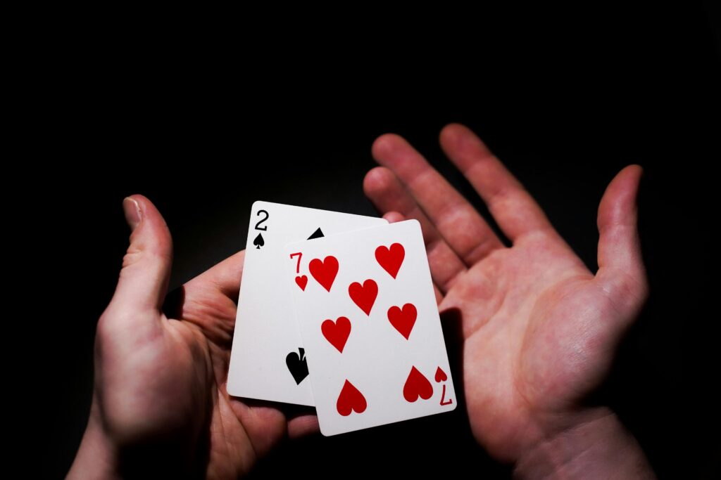 A person holds two playing cards, the two of spades and the seven of hearts, in their left hand with the right hand open and empty against a dark background. | FintechZoom