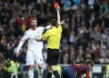 A soccer referee holds up a red card to a Real Madrid player as a Barcelona player kneels on the field. The Real Madrid player looks surprised, and a crowd watches in the background. | FintechZoom