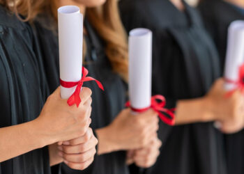 Four graduates in black gowns hold rolled diplomas tied with red ribbons, standing side by side. Their faces are not visible; the focus is on their hands and the diplomas. | FintechZoom