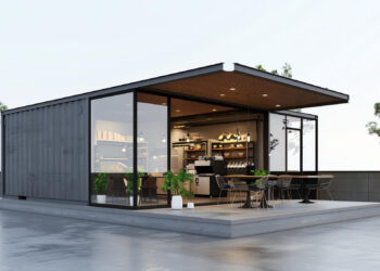 A modern Shipping Container Restaurant coffee shop featuring large glass walls, outdoor seating with black chairs and tables, potted plants, and a minimalist rooftop setting with a few small trees. | FintechZoom
