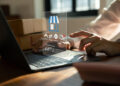 A person using a laptop with virtual icons for online shopping, including options for review, return, cancellation, and tracking shipment, displayed above the keyboard. Cardboard boxes are visible in the background. | FintechZoom