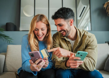 A smiling couple sits on a couch, looking at a smartphone together. The woman holds a credit card while the man points at her phone and holds another phone, suggesting online shopping or a digital transaction. | FintechZoom