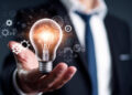A person in a suit holds a glowing light bulb, with gear and network icons surrounding it, symbolizing innovation, ideas, and technology in business. | FintechZoom