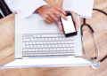 A person wearing a white lab coat uses a smartphone while resting it on a laptop keyboard. A stethoscope lies on the wooden desk next to the laptop. | FintechZoom