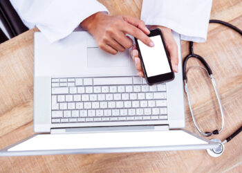 A person wearing a white lab coat uses a smartphone while resting it on a laptop keyboard. A stethoscope lies on the wooden desk next to the laptop. | FintechZoom