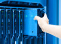 A person’s hand installs or removes a server blade into a rack of servers in a data center, with blue lighting illuminating the equipment. | FintechZoom