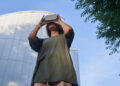 A person wearing a green shirt uses a virtual reality headset outdoors, standing in front of a modern, curved glass building with trees framing the scene against a blue sky. | FintechZoom