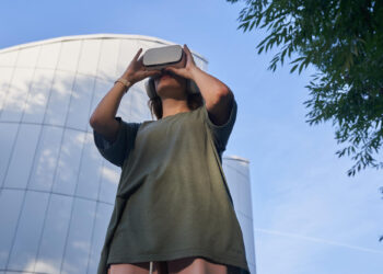A person wearing a green shirt uses a virtual reality headset outdoors, standing in front of a modern, curved glass building with trees framing the scene against a blue sky. | FintechZoom