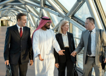 Four business professionals, three men and one woman, walk together through a modern glass walkway, engaged in conversation. One man wears a traditional white thawb and ghutra, while the others wear business suits. | FintechZoom