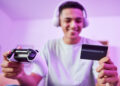 A young person wearing headphones smiles while holding a game controller in one hand and a credit card in the other, suggesting online gaming or digital purchases, with a purple-lit background. | FintechZoom