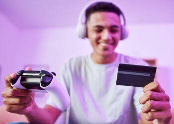 A young person wearing headphones smiles while holding a game controller in one hand and a credit card in the other, suggesting online gaming or digital purchases, with a purple-lit background. | FintechZoom