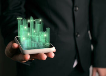 A person in a suit holds a smartphone displaying a green, holographic cityscape with glowing buildings and circular lights, symbolizing futuristic technology and smart city concepts. | FintechZoom