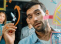 A man writes on a transparent board with a marker while two colleagues look on, surrounded by colorful notes and diagrams. The group appears to be brainstorming or planning together. | FintechZoom