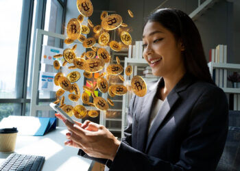A woman in business attire smiles while using her smartphone at a desk. Illustrated Bitcoin coins appear to burst from her phone, suggesting digital currency success. A coffee cup and computer are on the desk. | FintechZoom