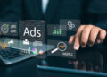 A person uses a laptop with floating digital icons related to online advertising, such as “Ads,” magnifying glass, graph, target, and gears, symbolizing digital marketing and analytics. | FintechZoom