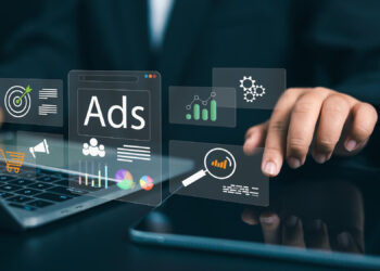 A person uses a laptop with floating digital icons related to online advertising, such as “Ads,” magnifying glass, graph, target, and gears, symbolizing digital marketing and analytics. | FintechZoom
